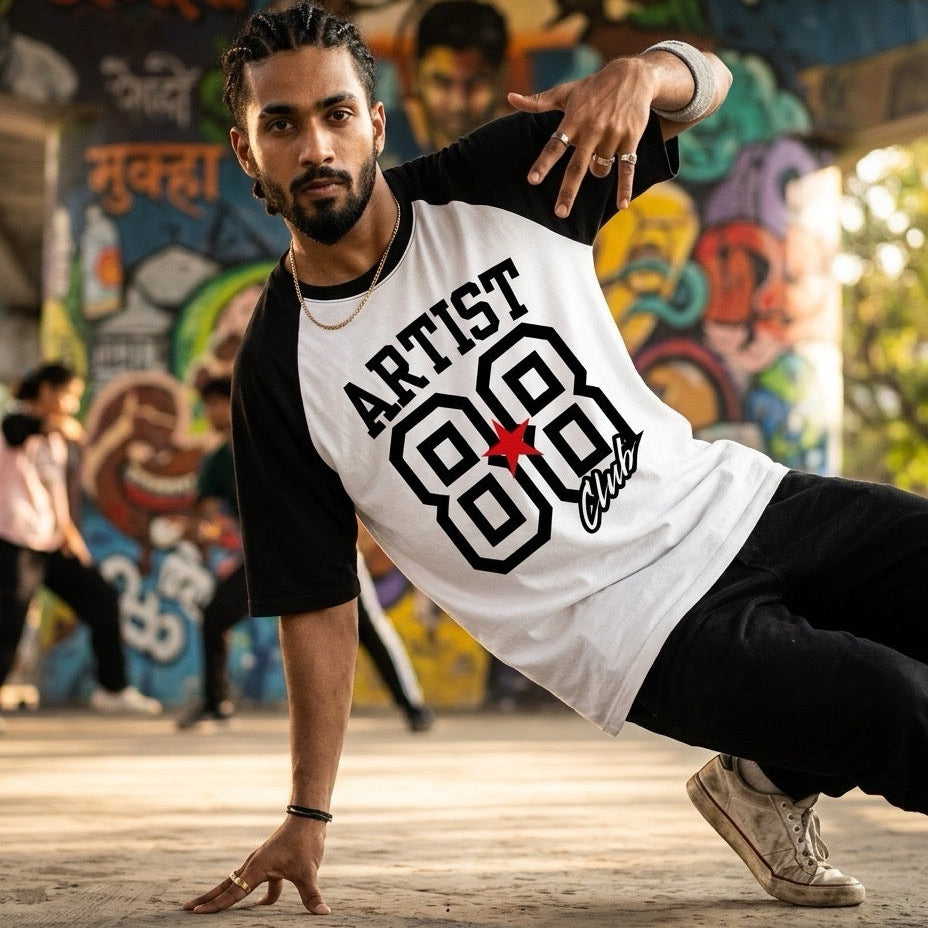 A man in a black and white raglan t-shirt with 'ARTIST Club 88' printed on the front, posing in a dynamic handstand pose in front of a colorful graffiti wall.