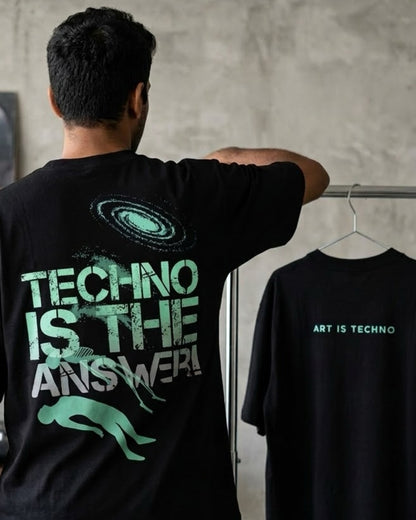 A DJ stands with his back to the camera wearing a black t-shirt with a large green and white graphic on the back that reads 'TECHNO IS THE ANSWER!'. To his right, a black t-shirt on a hanger displays the text 'ART IS TECHNO' in green and white against a textured gray wall.