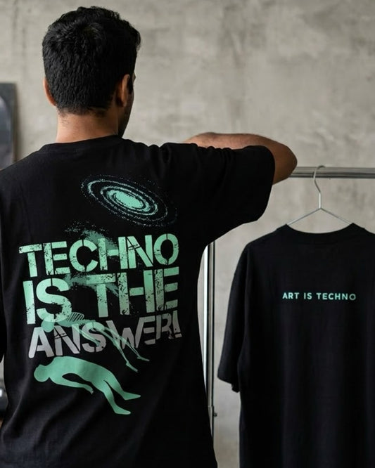 A DJ stands with his back to the camera wearing a black t-shirt with a large green and white graphic on the back that reads 'TECHNO IS THE ANSWER!'. To his right, a black t-shirt on a hanger displays the text 'ART IS TECHNO' in green and white against a textured gray wall.