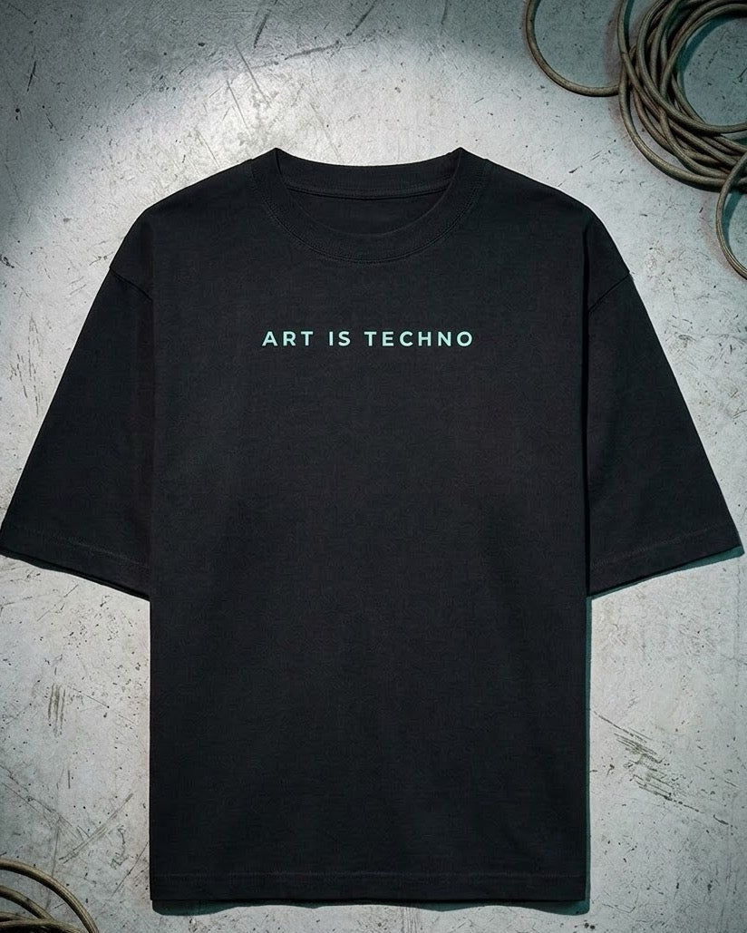 A black short-sleeve t-shirt with the text 'ART IS TECHNO' printed in a light teal-green font across the chest, displayed flat on a textured gray concrete surface with coiled cables in the corners.