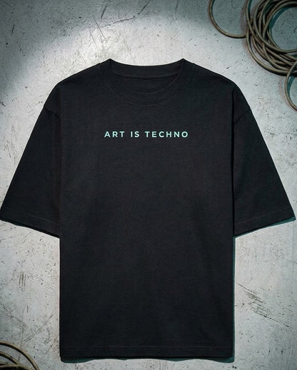 A black short-sleeve t-shirt with the text 'ART IS TECHNO' printed in a light teal-green font across the chest, displayed flat on a textured gray concrete surface with coiled cables in the corners.