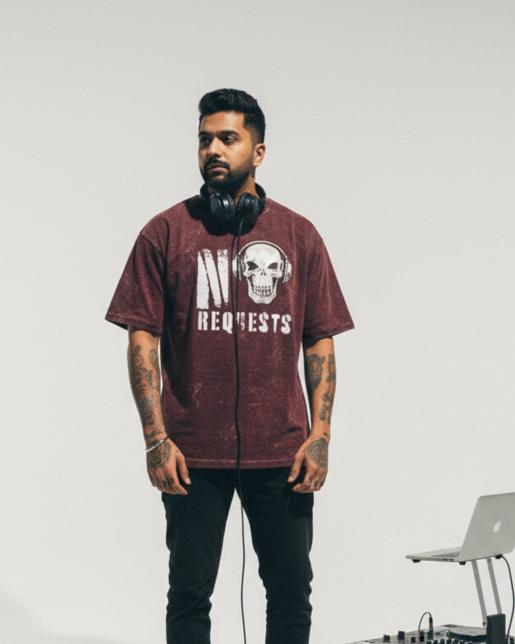 Man wearing a maroon t-shirt with a skull design and text, standing next to DJ equipment on a plain background