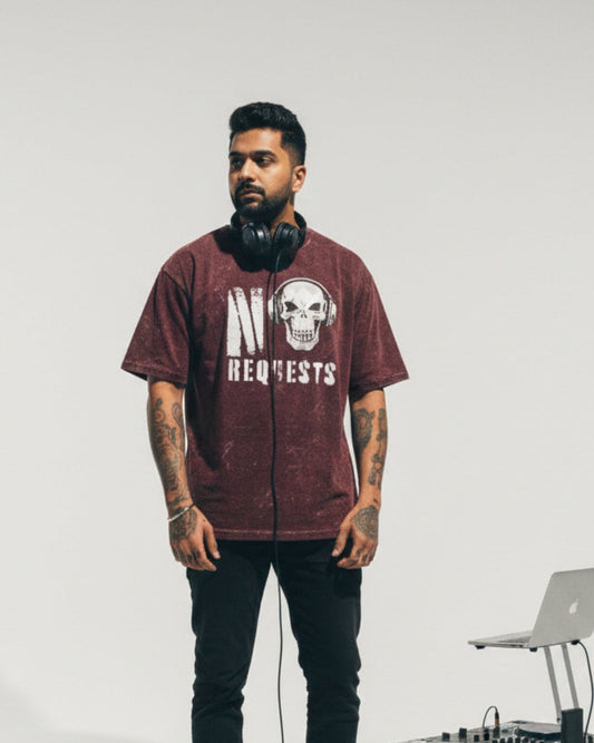 Man wearing a maroon t-shirt with a skull design and text, standing next to DJ equipment on a plain background