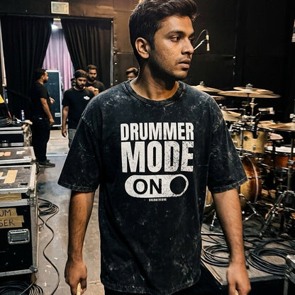 Person wearing a 'DRUMMER MODE ON' t-shirt in a drumming studio.