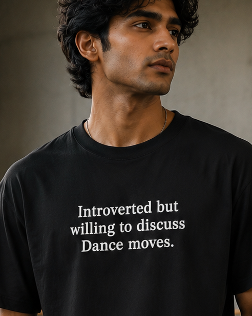 A man wearing a black DrummerKong oversized t-shirt with white text that reads 'Introverted but willing to discuss Dance moves.' and black sweatpants, standing in a concrete studio with ballet barre.