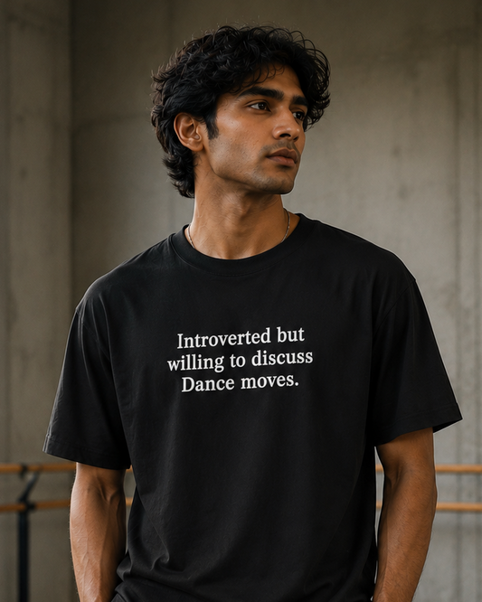 A man wearing a black DrummerKong oversized t-shirt with white text that reads 'Introverted but willing to discuss Dance moves.' and black sweatpants, standing in a concrete studio with ballet barre.
