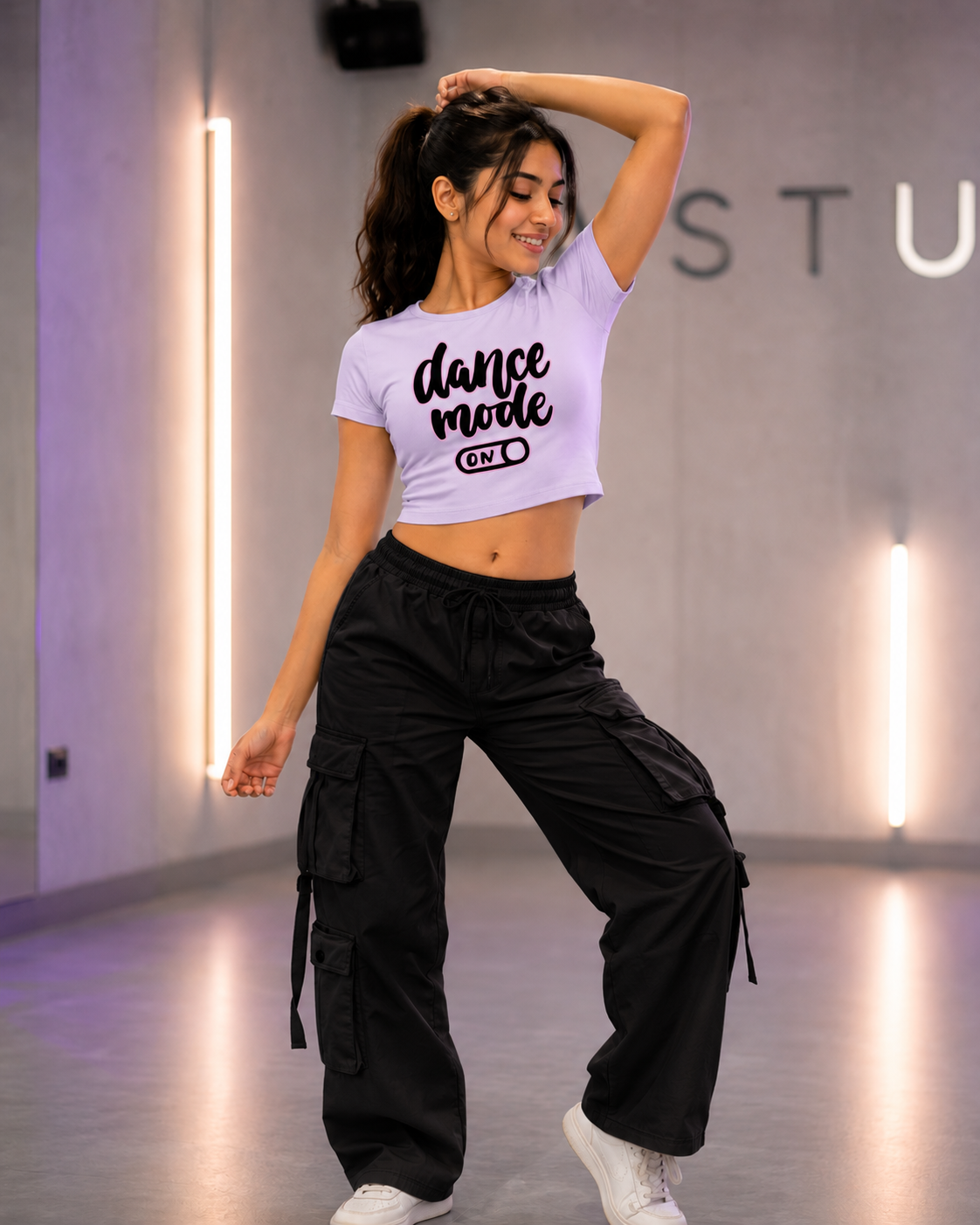 A dancer posing in a dance studio wearing a light purple crop top with 'dance mode ON' printed on the front, paired with black cargo pants and white sneakers.
