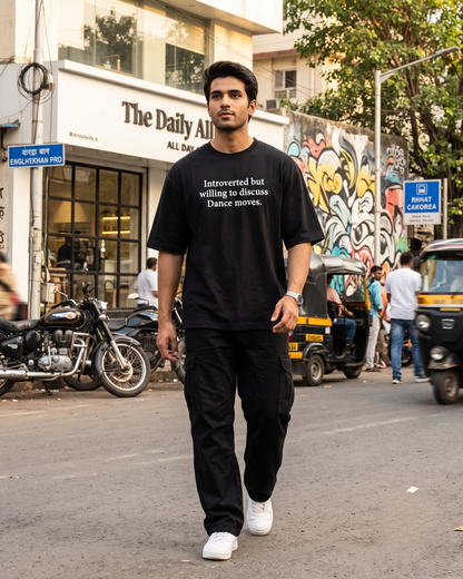 A dancer walking down a city street wearing a DrummerKong oversized t-shirt with white text, black cargo pants, and white sneakers.