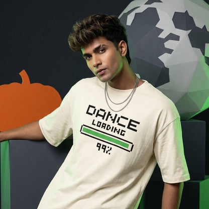 A man wearing a DrummerKong's cream-colored graphic t-shirt with 'DANCE LOADING 99%' printed on the front, paired with black pants, posed in front of a dark background with geometric pumpkin and globe decorations.