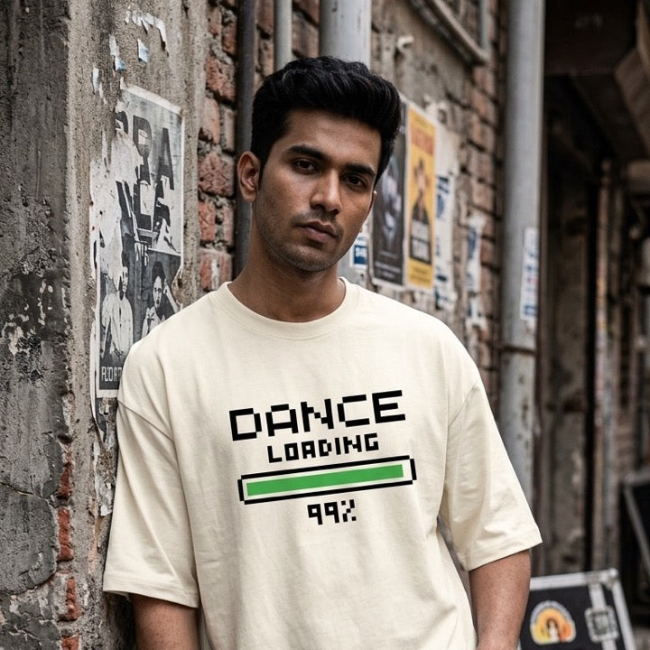 A dancer wearing a cream-colored graphic t-shirt with 'DANCE LOADING 99%' printed on the front, leaning against a weathered brick wall in an alley.