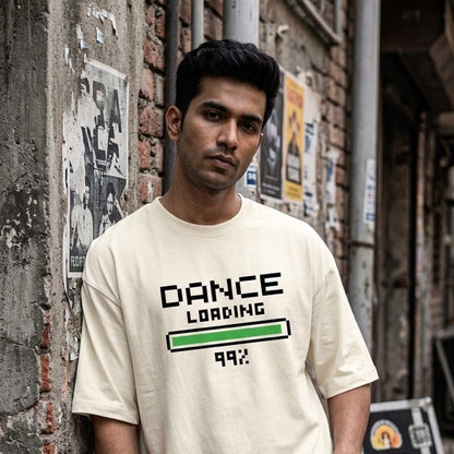A dancer wearing a cream-colored graphic t-shirt with 'DANCE LOADING 99%' printed on the front, leaning against a weathered brick wall in an alley.