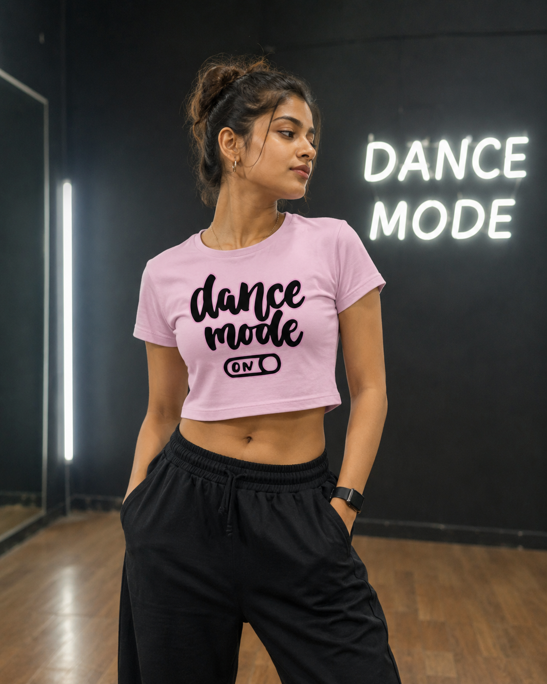 A girl dancer posing in a dance studio wearing a light pink crop top with a black graphic that reads 'dance mode ON ' paired with black sweatpants.