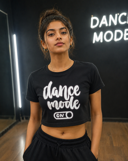 A girl dancer wearing a drummerkong's black cropped t-shirt with 'dance mode ON' printed in white script, standing in a dance studio with a black wall and a neon sign in the background.