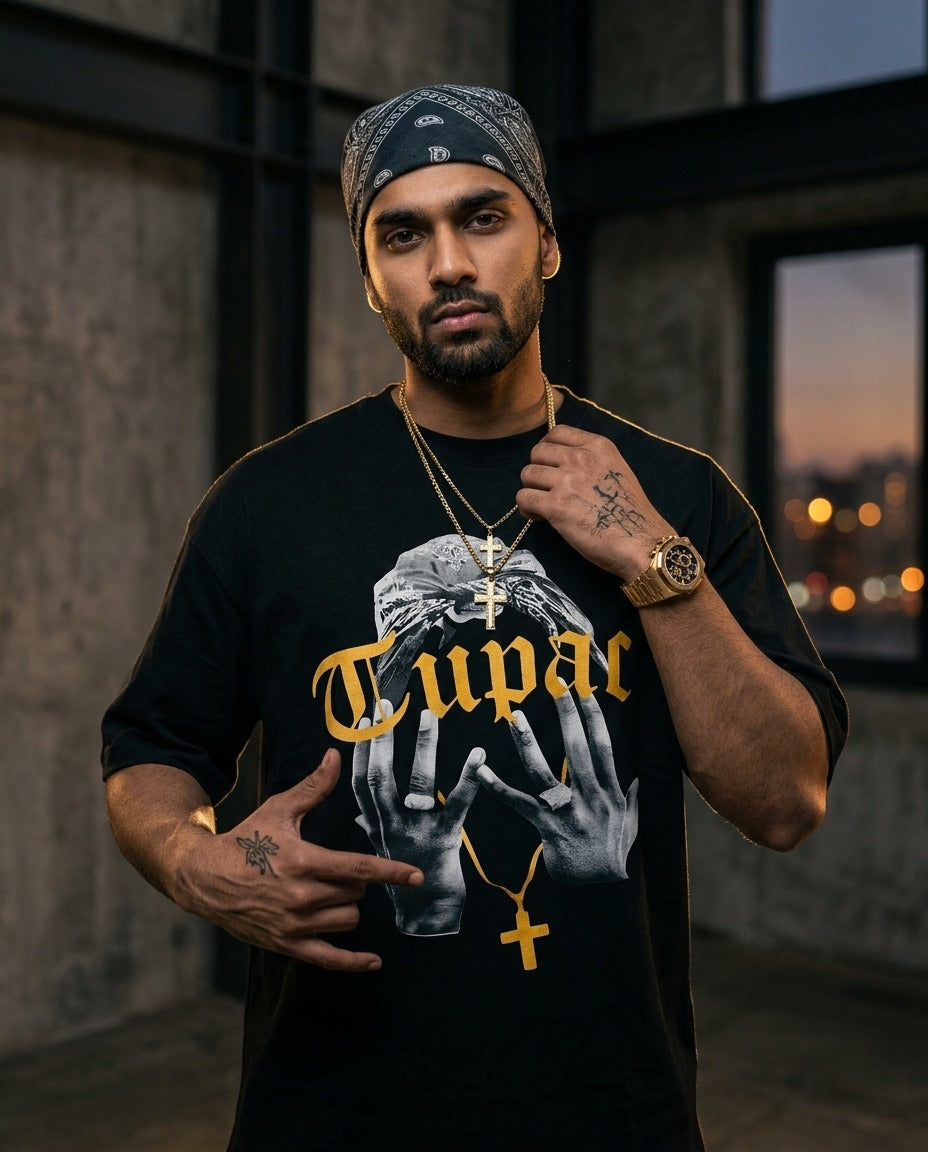 Man wearing a black T-shirt with 'Tupac' design and gold text, standing in an urban setting.